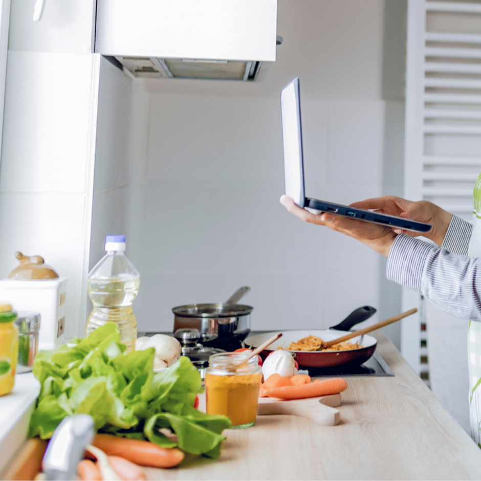 Person am Kochen mit Gemüse und einem Laptop in der Hand in der Küche.