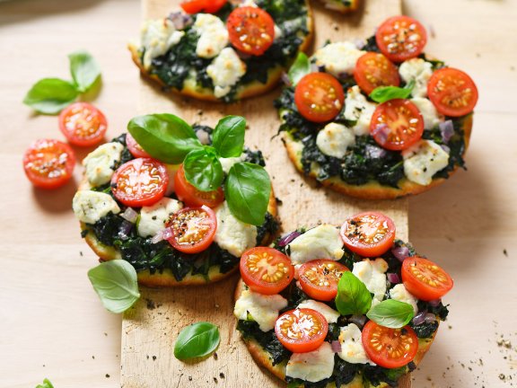 Mit Tomaten und Ziegenkäse belegte und gebackene Minipizzen auf einem Holzbrett.